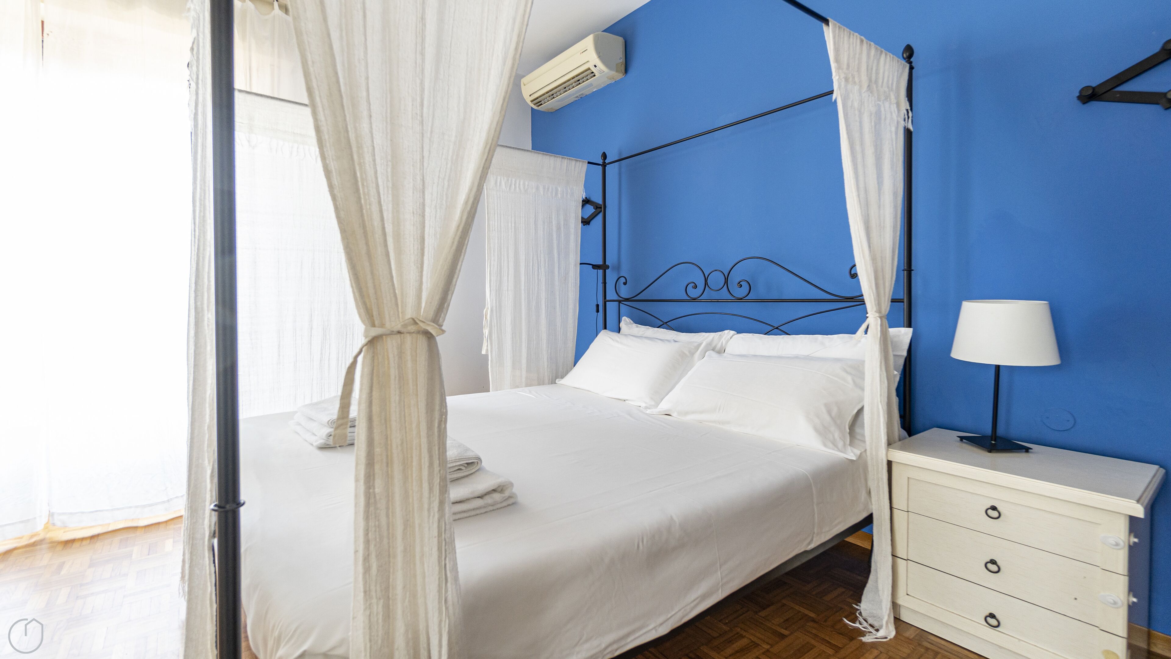A bedroom with a bed, white curtains, a bedside table with a lamp, and a blue wall.
