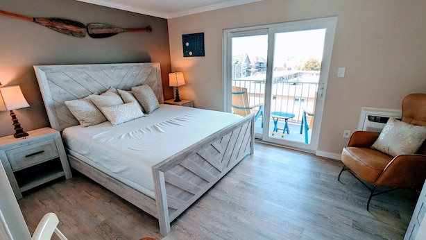 Ein Schlafzimmer mit Bett, Nachttischen, einem Sessel, Holzboden und Blick durch eine Glastür nach draußen.