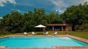 A rectangular swimming pool with a stone border, surrounded by a grassy area with four lounge chairs and a white umbrella.