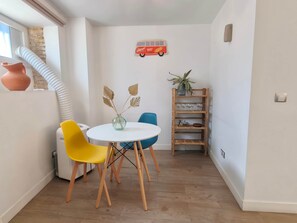 Un pequeño comedor con una mesa redonda, dos sillas, un jarrón con hojas decorativas, un adorno de autobús rojo en la pared, una estantería de madera con vasos y una bicicleta, y una planta en maceta.