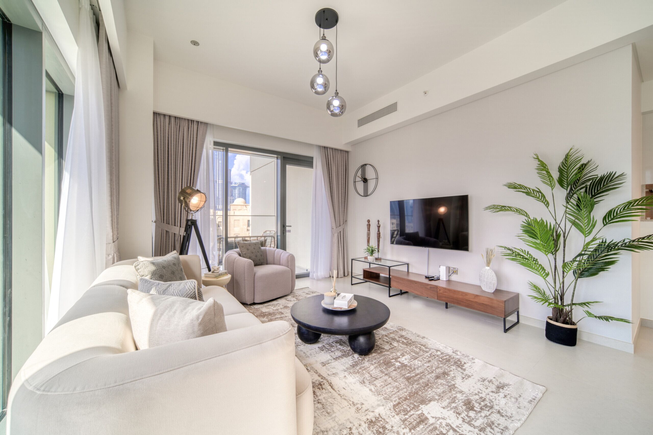 Una sala de estar moderna con un sofá, una mesa de centro y un televisor. Hay una ventana grande con cortinas, una planta y un reloj en la pared.