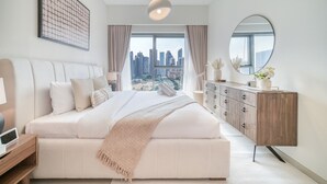 A modern bedroom with a large bed, a wooden dresser, a round mirror, and a city view.