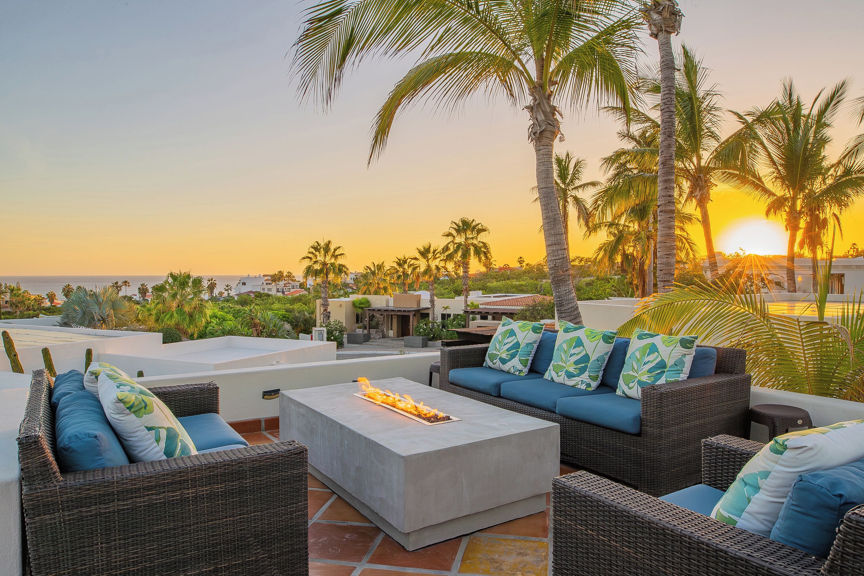 A rooftop patio with wicker furniture, a fire feature, and a view of the sunset over the ocean.