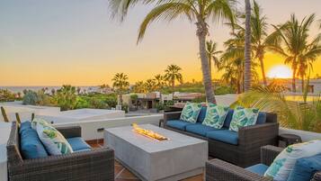 A rooftop patio with wicker furniture, a fire feature, and a view of the sunset over the ocean.