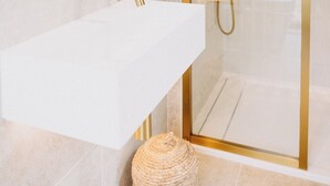 Un baño moderno con un lavabo blanco, detalles dorados, una ducha con cerramiento de vidrio y una planta en el mueble.