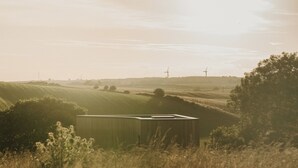 Paisaje rural con colinas onduladas, un aerogenerador y un pequeño edificio en primer plano.