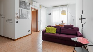 Un salon avec un canapé violet, une petite table et une lampe. Il y a une porte qui donne sur une autre pièce et une fenêtre avec des rideaux.