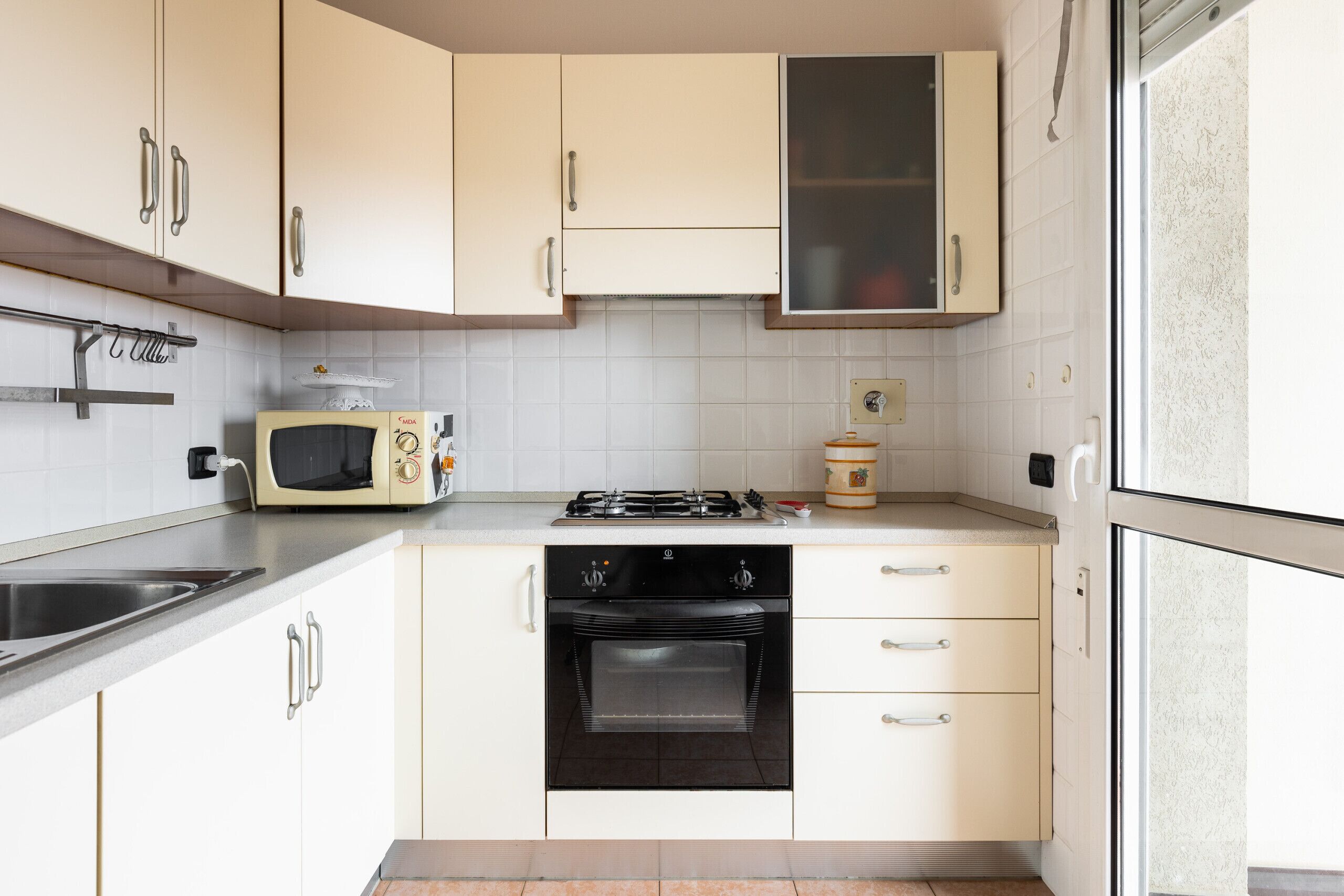 Une cuisine compacte avec des meubles blancs, un four à micro-ondes encastré, une cuisinière à gaz et un évier en acier inoxydable.