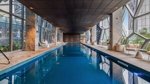 A modern indoor swimming pool with clear glass walls, lounge seating, and a geometric ceiling design. - CBD, Stylish, Heated Pool, Gym, Sauna, Steam, BBQ (Melbourne)