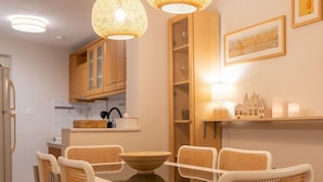Un comedor con una mesa blanca y sillas de mimbre, dos lámparas colgantes de mimbre, una cocina al fondo y una alfombra a rayas.