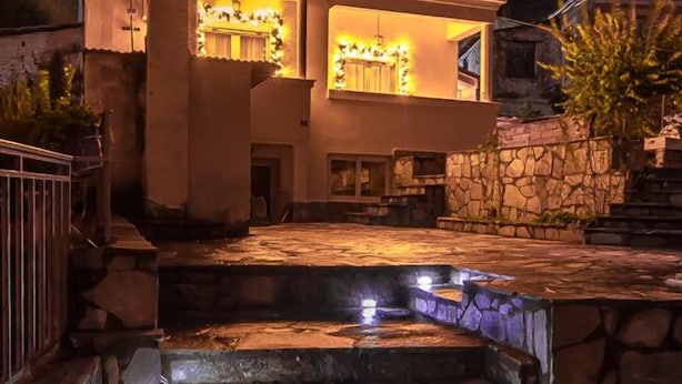 Una casa moderna con fachada de piedra, una escalera de piedra que lleva a una piscina y una gran formación rocosa al fondo.