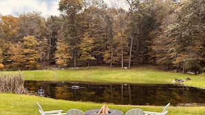 A serene outdoor seating area with Adirondack chairs and a fire pit, overlooking a pond and surrounded by autumn foliage. - The Cozy Goshen Home, 5mins from LEGOLAND and near skiing! (Goshen)