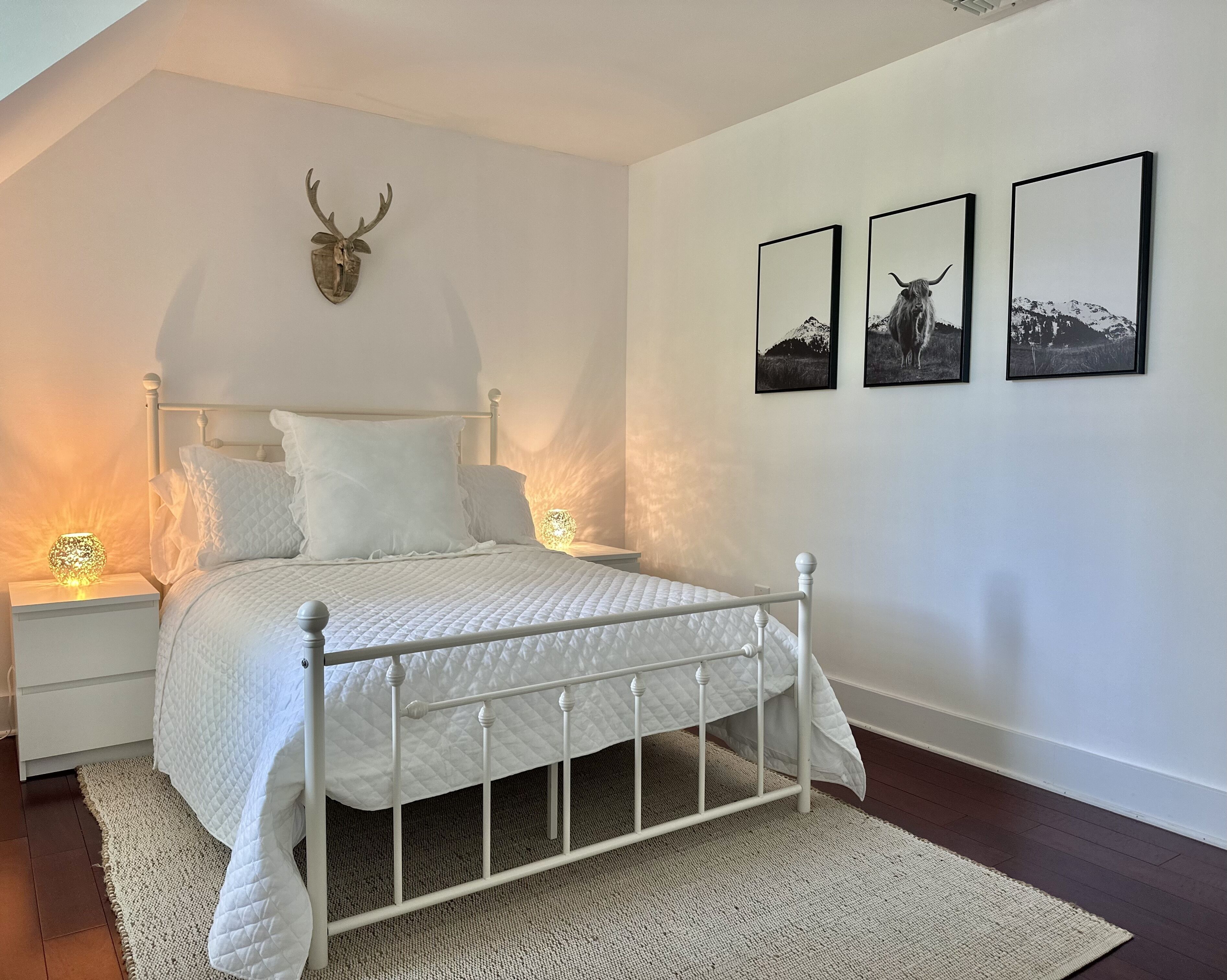 Un dormitorio con una cama blanca, una cabeza de venado decorativa en la pared y tres cuadros enmarcados.