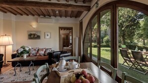 A cozy living room with a wooden ceiling, a sofa, a dining table with chairs, and a large window overlooking a garden.