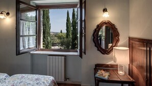 A bedroom with an open window, a wooden dresser, a mirror, and a bedside table with a lamp and a book.
