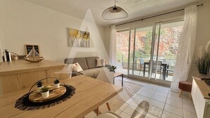 A modern dining area with a wooden table, white chairs, and a black woven placemat. There is a sofa and a dining table set in the background. - T3 sur les hauteurs dans résidence avec piscine (Sète)