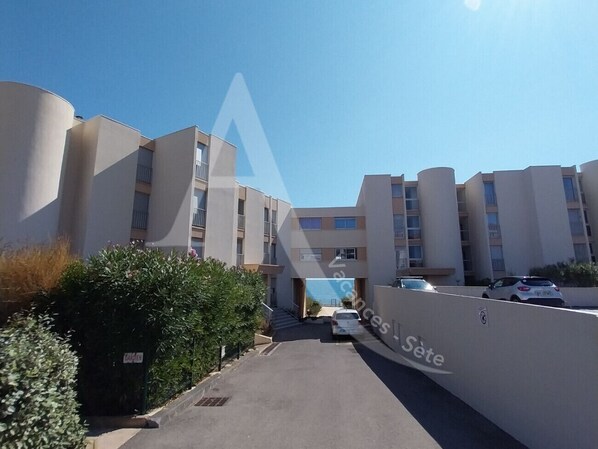 A multi-story residential building with a central entrance, a parking area, and a clear sky.