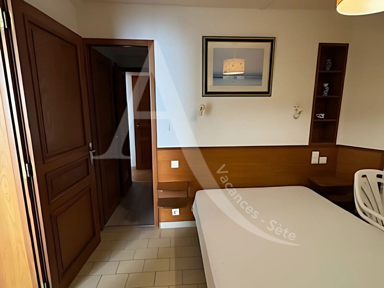 A hotel room with a bed, a wooden wardrobe, a framed picture on the wall, and a small shelf with items.