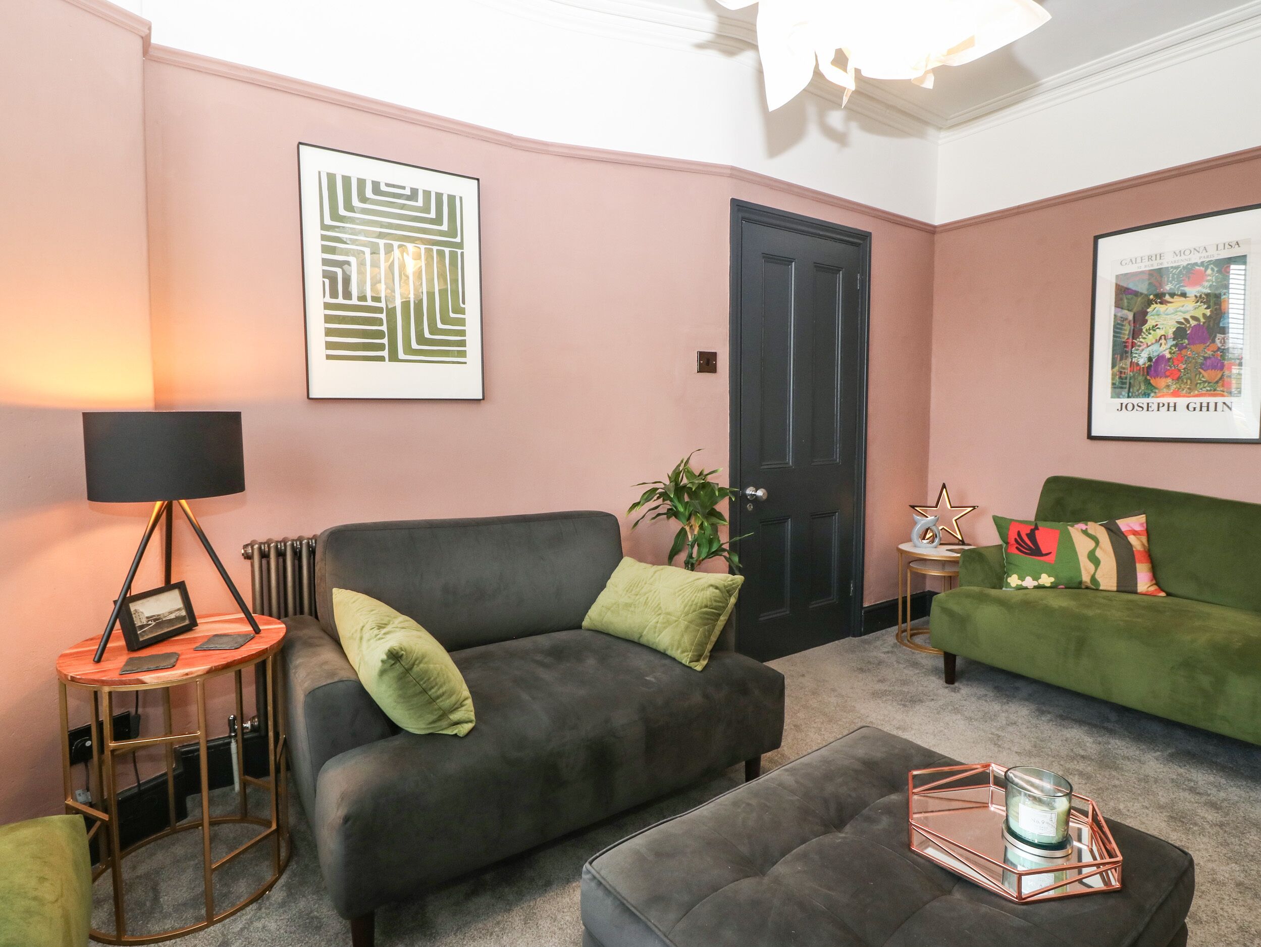 Un salon avec un canapé gris, un canapé vert, un fauteuil noir, une petite table ronde avec une lampe et un tableau encadré au mur.