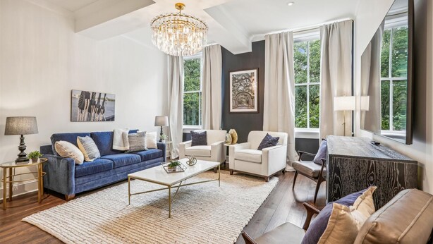 A spacious living room with a chandelier, a blue sofa, a white sofa, and a coffee table. There are large windows with curtains, a painting on the wall, and a lamp on a side table. - NEW REMODEL Chalmers at the Historic Bryson Building (Savannah)