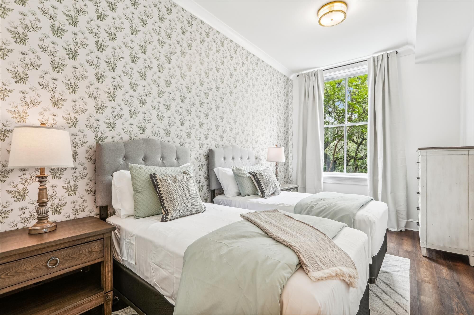 Une chambre avec deux lits, un papier peint à motifs floraux, une fenêtre avec des rideaux et une table de chevet avec une lampe.