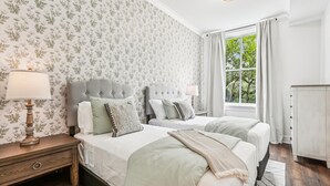 A bedroom with two beds, floral wallpaper, a window with curtains, and a nightstand with a lamp. - NEW REMODEL Chalmers at the Historic Bryson Building (Savannah)