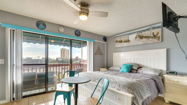 Ein Schlafzimmer mit Bett, Nachttisch, Tisch mit Stühlen, Deckenventilator, Fenster mit Blick auf Gebäude und einem Wandbild.