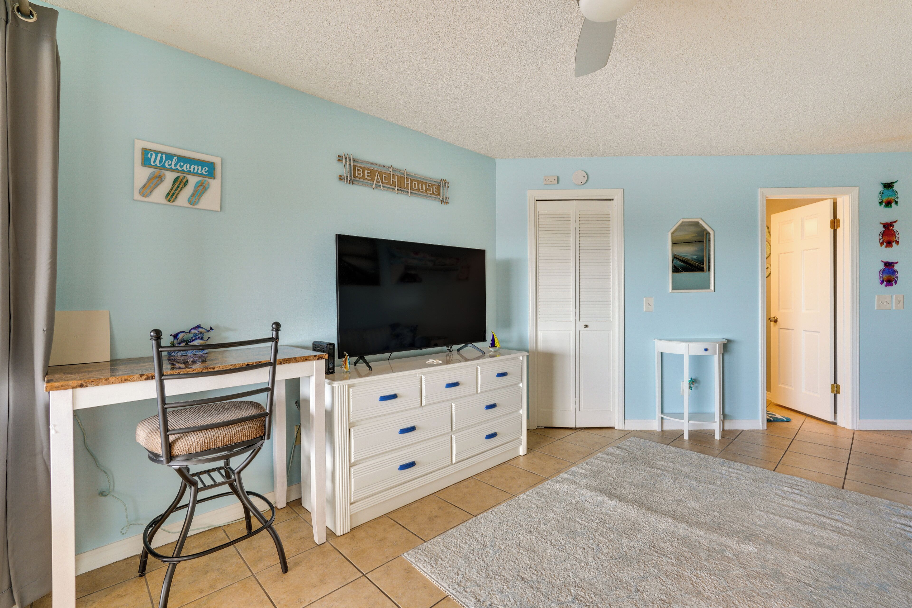 A room with a TV, a desk, a chair, a dresser, a door, and a wall sign.