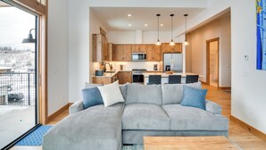 A modern living room with a grey sectional sofa, wooden flooring, and a view of a kitchen and dining area through an open door. - Winter Park Retreat: Balcony, Hot Tub & Fireplace! (Fraser)