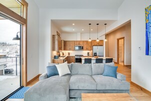 Una sala de estar moderna con un sofá seccional gris, piso de madera y vistas a una cocina y comedor a través de una puerta abierta.