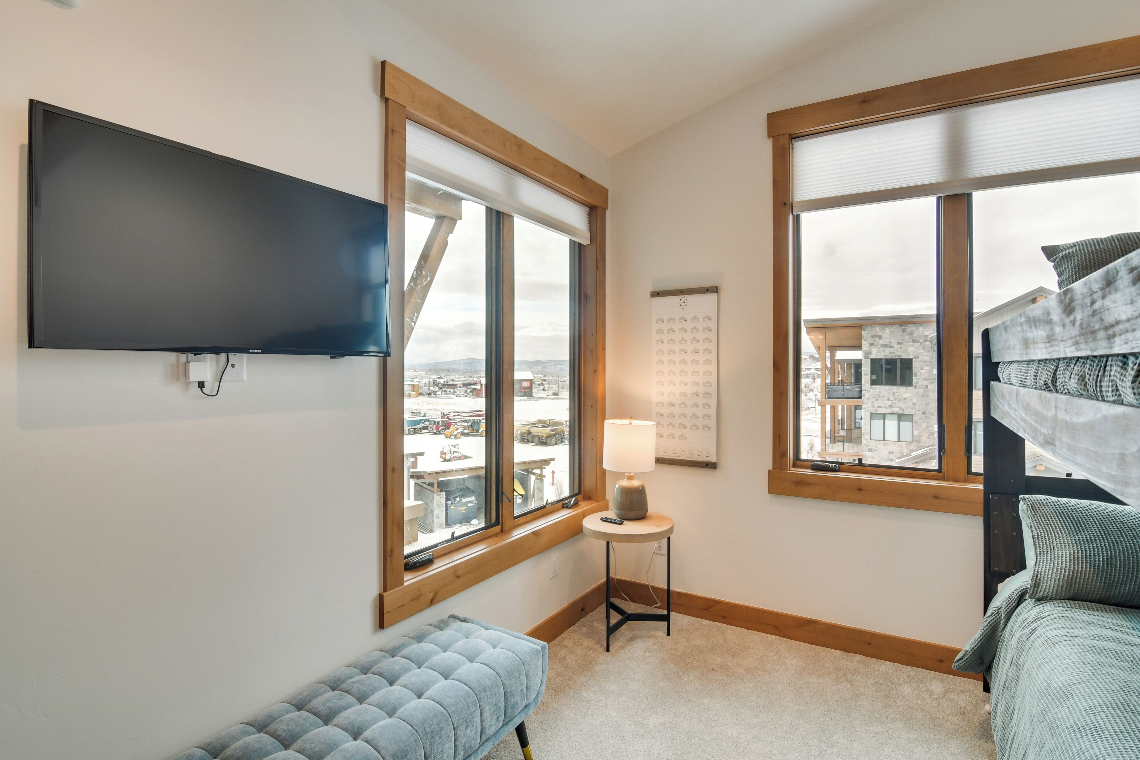 A bedroom with a large window, a flat-screen TV, a bedside table with a lamp, and a bed with a headboard.