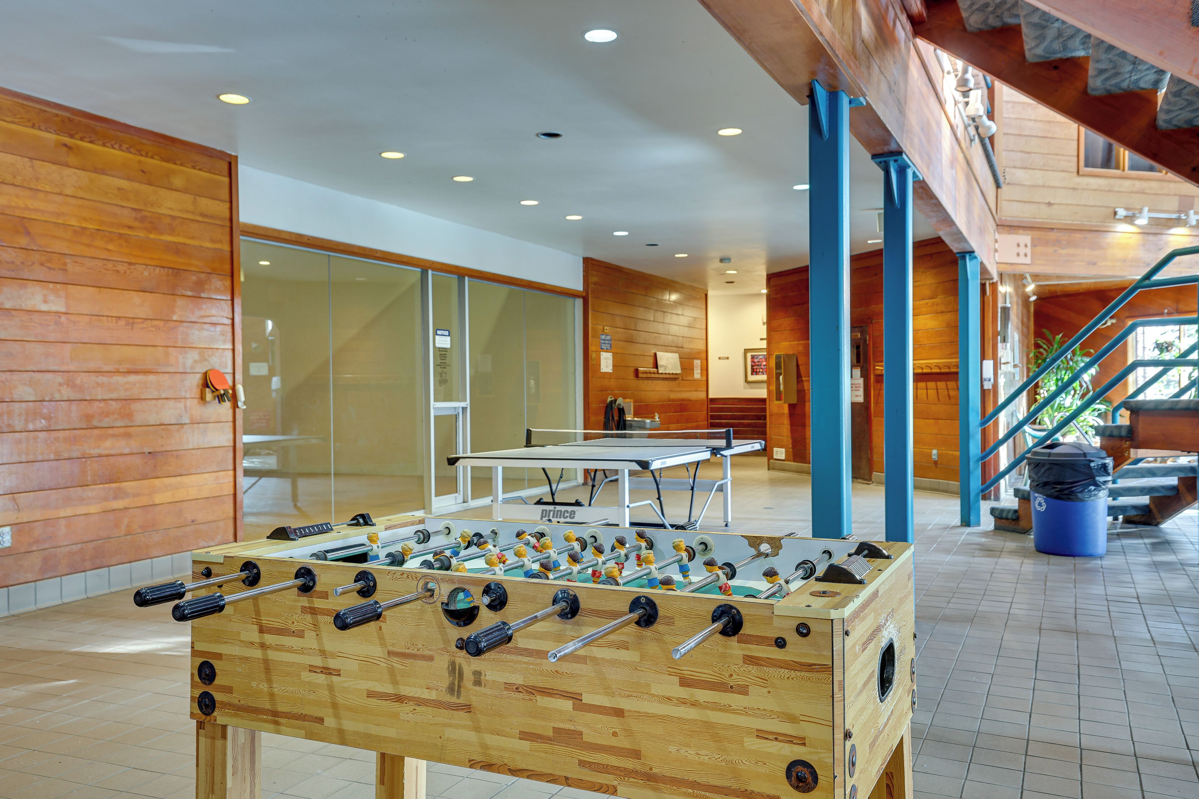 A foosball table in a lobby with wooden walls and a ping pong table in the background.