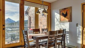Une table à manger en bois avec des chaises, une vue sur les montagnes par une porte vitrée et un tableau représentant un coq accroché au mur.