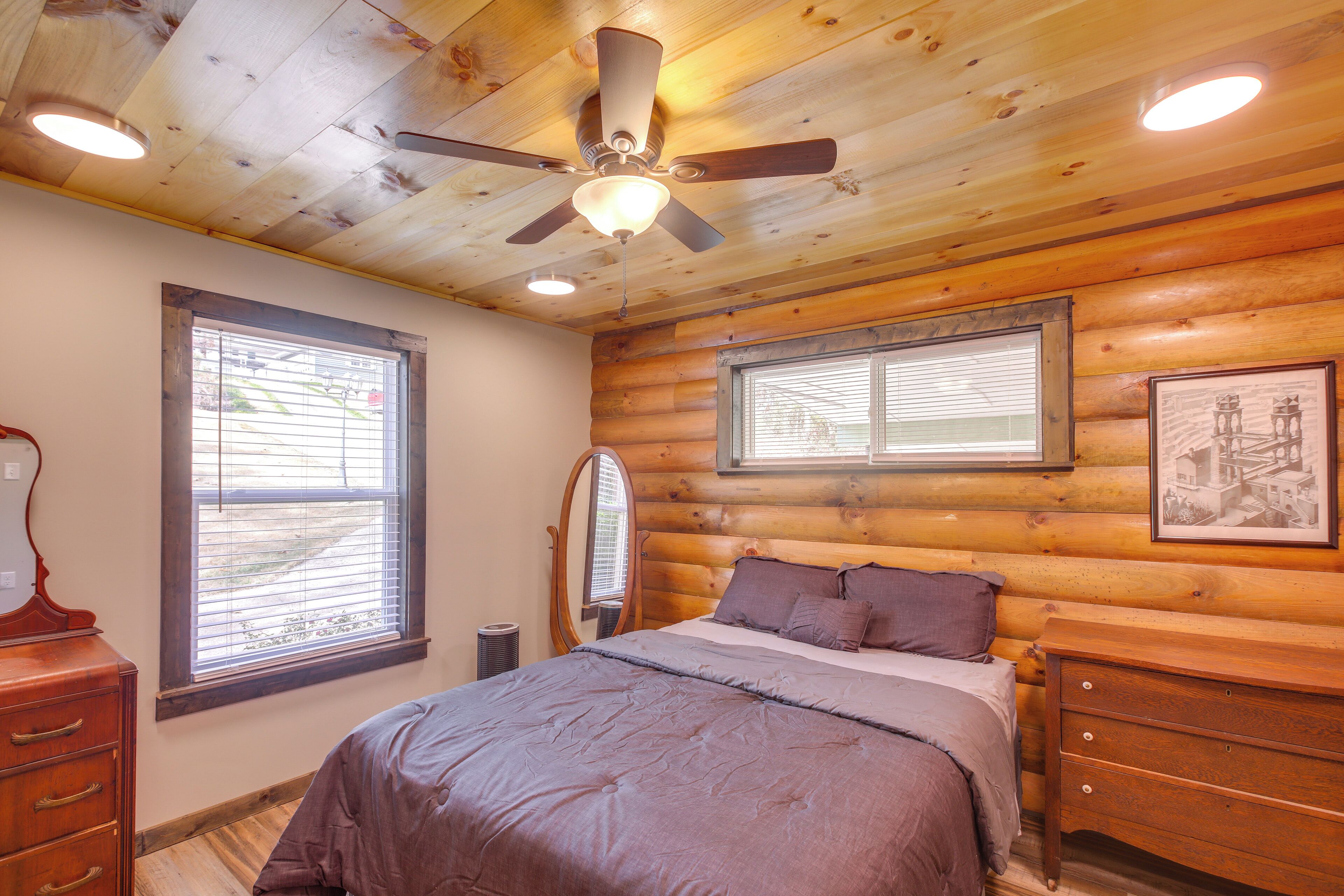 A bedroom with a bed, wooden walls, a ceiling fan, and two windows with blinds.