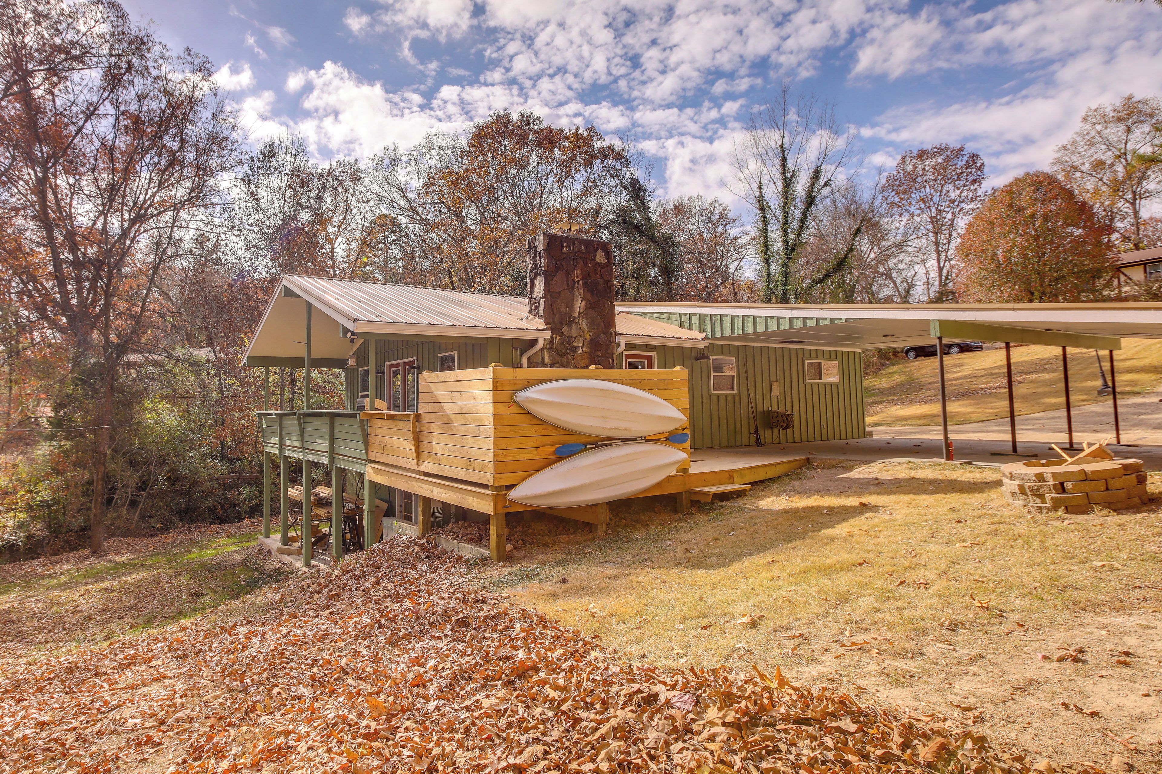 A rustic cabin with a wooden deck, a stone chimney, and a covered area with a car parked nearby.