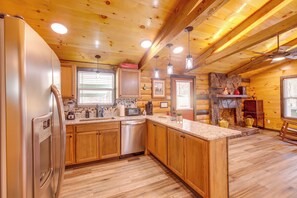 A log cabin kitchen with wooden cabinets, a stainless steel refrigerator, a dishwasher, a granite countertop, and a stone fireplace. - Pet-friendly Chattanooga Cabin w/ Hot Tub & Kayaks (Chattanooga)
