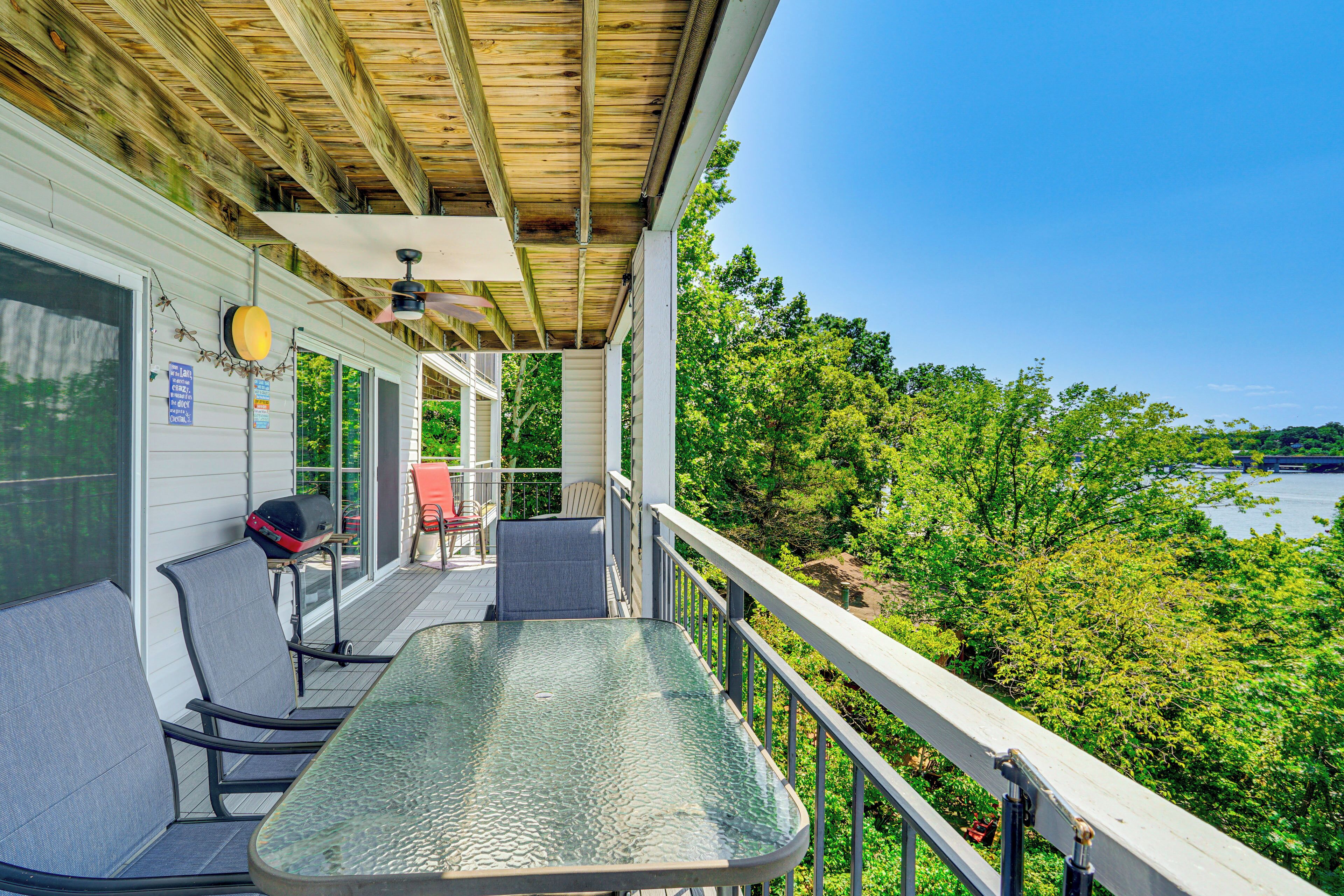 A covered outdoor area with a glass table and chairs, a barbecue grill, and a view of a lake surrounded by trees.