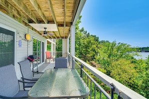 A covered outdoor area with a glass table and chairs, a barbecue grill, and a view of a lake surrounded by trees. - Waterfront Gem w/ Double-wide Balcony & Sunsets (Osage Beach)