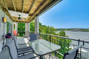 A covered outdoor deck with a table and chairs, overlooking a body of water and greenery. - Waterfront Gem w/ Double-wide Balcony & Sunsets (Osage Beach)