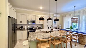 Eine moderne Küche mit weißen Schränken, einer Granit-Arbeitsplatte, einem Edelstahl-Kühlschrank, einem eingebauten Backofen, einer Mikrowelle und einer Kaffeemaschine. Es gibt drei Holzbarhocker und eine Hängeleuchte über der Kochinsel.