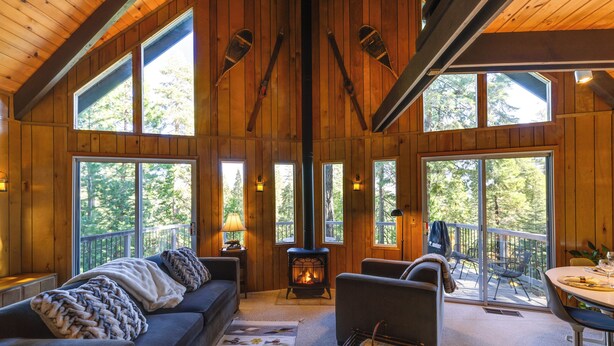 A cozy cabin interior with a fireplace, large windows, a sofa, and a dining area. - 3 Mi to Big Trees State Park: Cabin w/ Deck (Arnold)