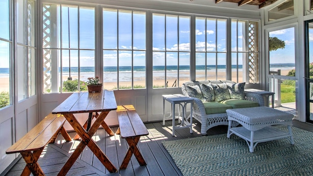 Une terrasse couverte avec une table et des bancs en bois, des meubles en osier blancs et de grandes fenêtres donnant sur la plage.