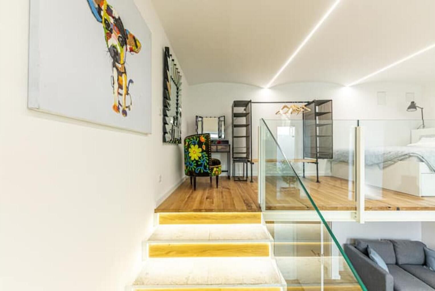 A modern interior with a staircase leading to a loft area, a glass railing, and a colorful chair.
