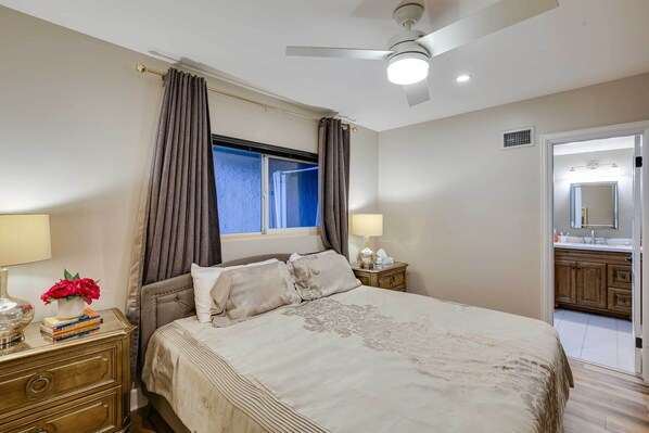 A bedroom with a bed, nightstands, a window with curtains, a ceiling fan, and a bathroom visible through an open door.