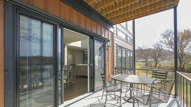 Un espacio al aire libre techado con una mesa y sillas, puertas correderas de vidrio que dan a un ambiente interior, y vista a un parque con árboles y colinas.