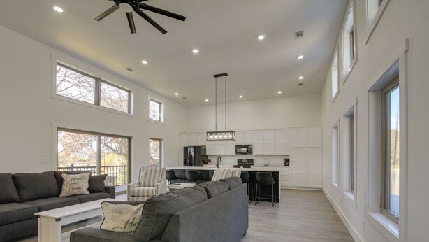 A spacious living room with a large gray sectional sofa, a white coffee table, and a dark gray couch. There is a kitchen area with white cabinets and a black countertop. The room has large windows and a ceiling fan. - Pet-friendly Cotter Retreat w/ Deck & Gas Grill! (Cotter)