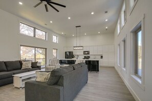 A spacious living room with a large gray sectional sofa, a white coffee table, and a dark gray couch. There is a kitchen area with white cabinets and a black countertop. The room has large windows and a ceiling fan. - Pet-friendly Cotter Retreat w/ Deck & Gas Grill! (Cotter)