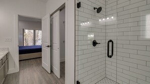 A modern bathroom with white subway tiles, a glass shower enclosure, and a visible door leading to a bedroom with a bed and a window. - Pet-friendly Cotter Retreat w/ Deck & Gas Grill! (Cotter)