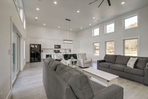 A spacious living room with a grey sectional sofa, a coffee table, and a striped armchair. A kitchen with white cabinets and a dining area with a table and chairs is visible in the background.