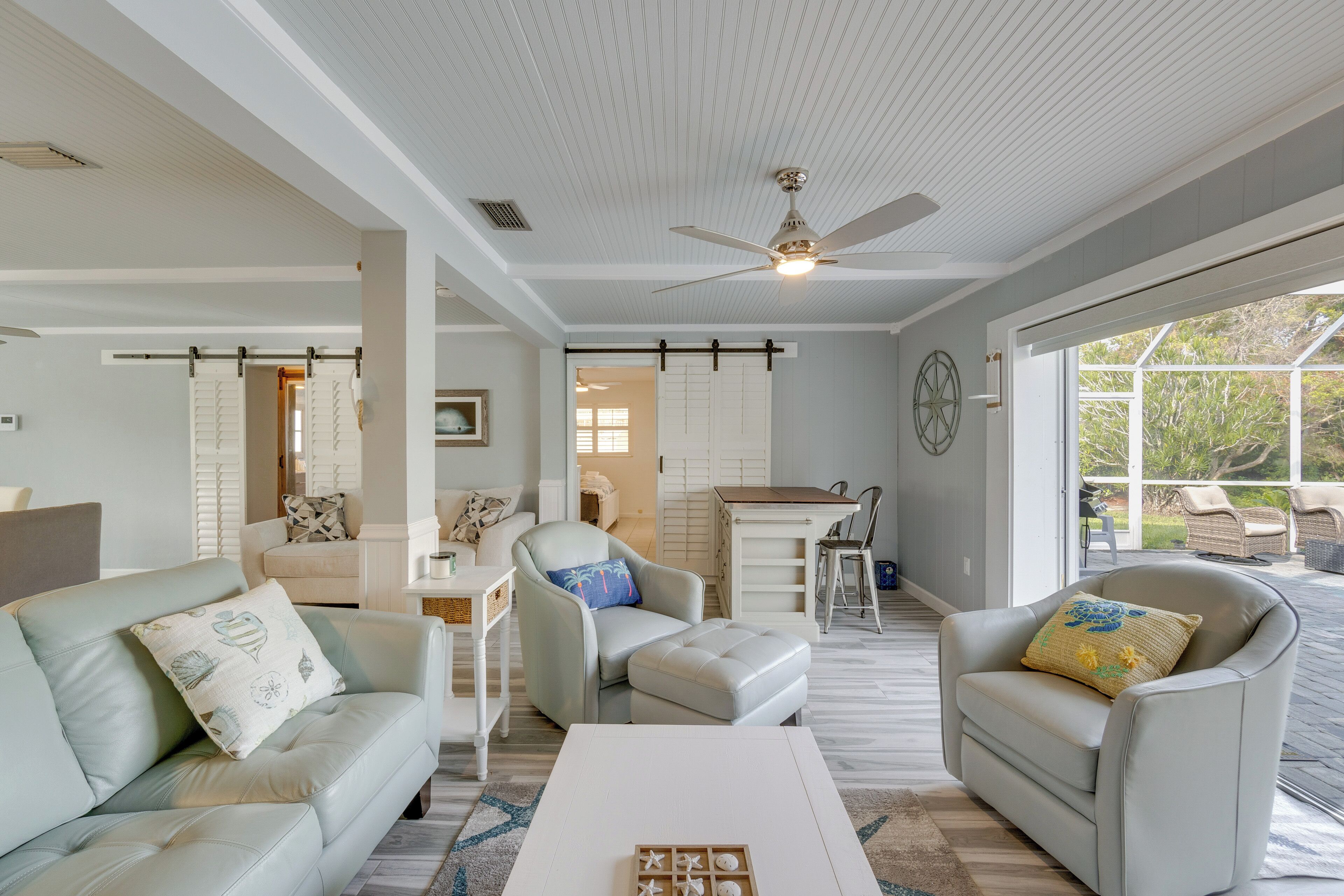 A spacious living area with a ceiling fan, a dining area with a table and chairs, and a view through large windows.